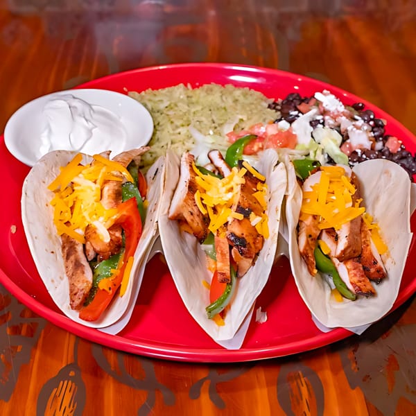 Three chicken fajita tacos with cheese, peppers, rice, black beans, and sour cream on a red plate.