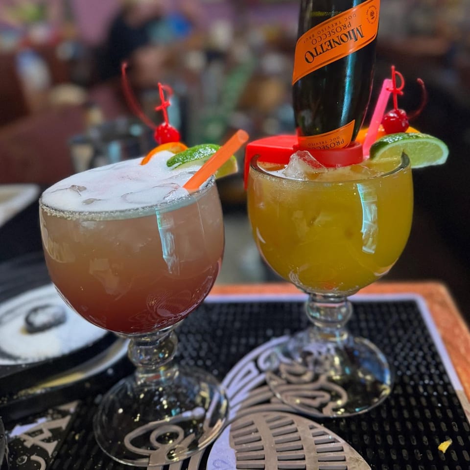 Two colorful Casa Vallarta cocktails, one with a mini Prosecco bottle, garnished with fruit and straws.