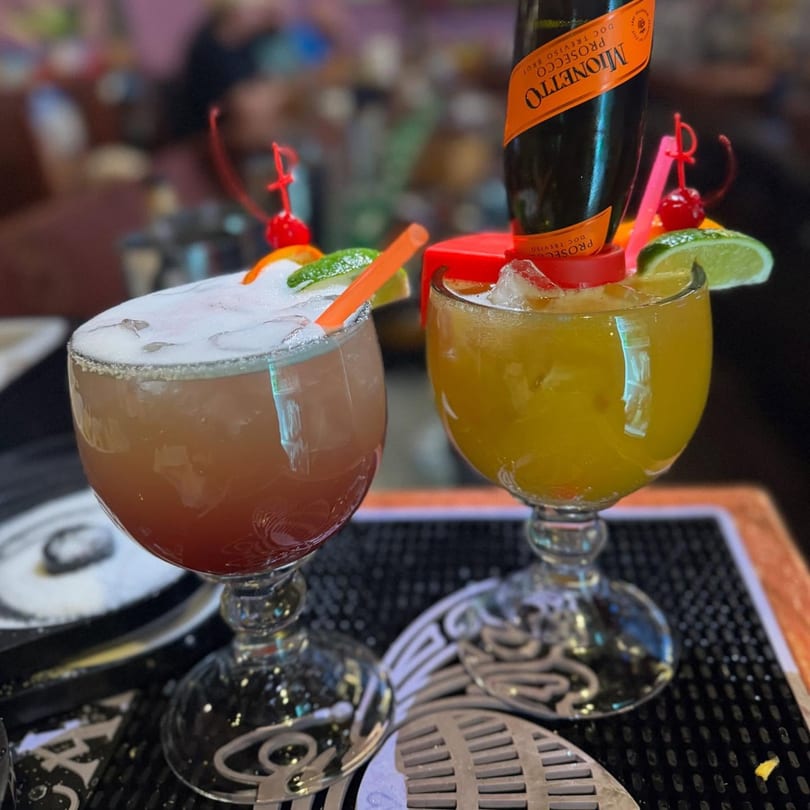 Two colorful Casa Vallarta cocktails, one with a mini Prosecco bottle, garnished with fruit and straws.