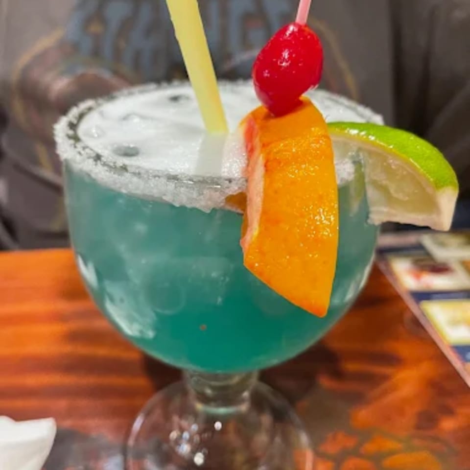 A vibrant blue margarita with a salted rim, garnished with orange, lime, and a cherry.
