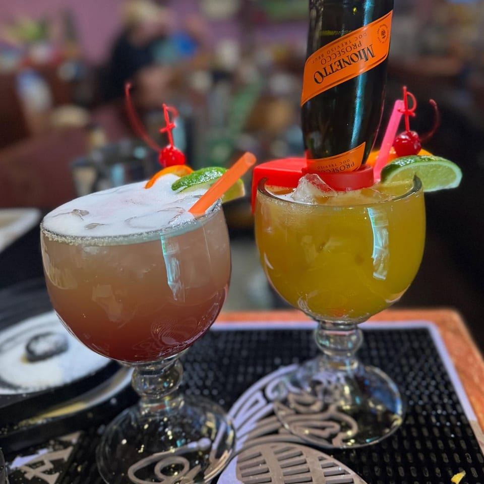 Two colorful Casa Vallarta cocktails, one with a mini Prosecco bottle, garnished with fruit on a bar.