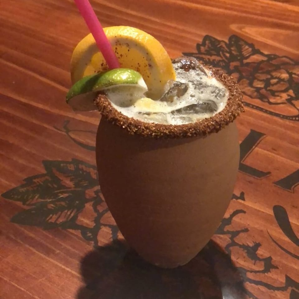 A Casa Vallarta Mexican cocktail in a clay cup with a spiced rim, lime, orange, and pink straw.