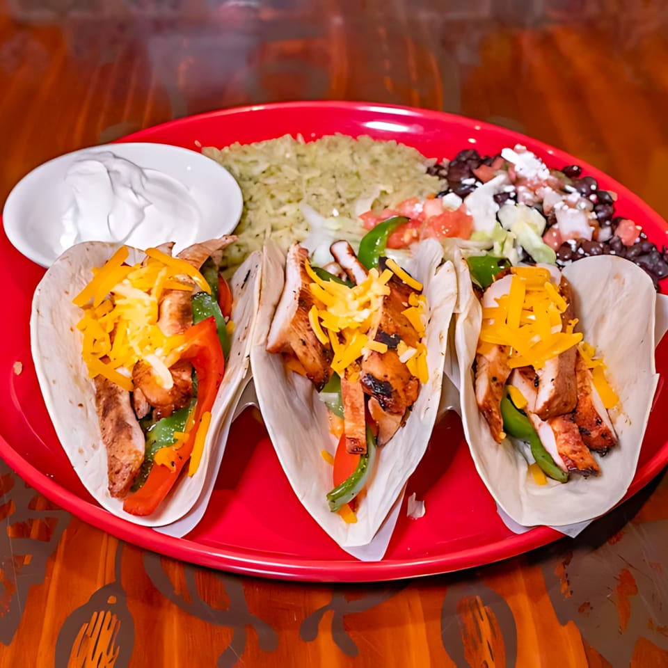 Three chicken fajita tacos with cheese, peppers, rice, black beans, and sour cream on a red plate.