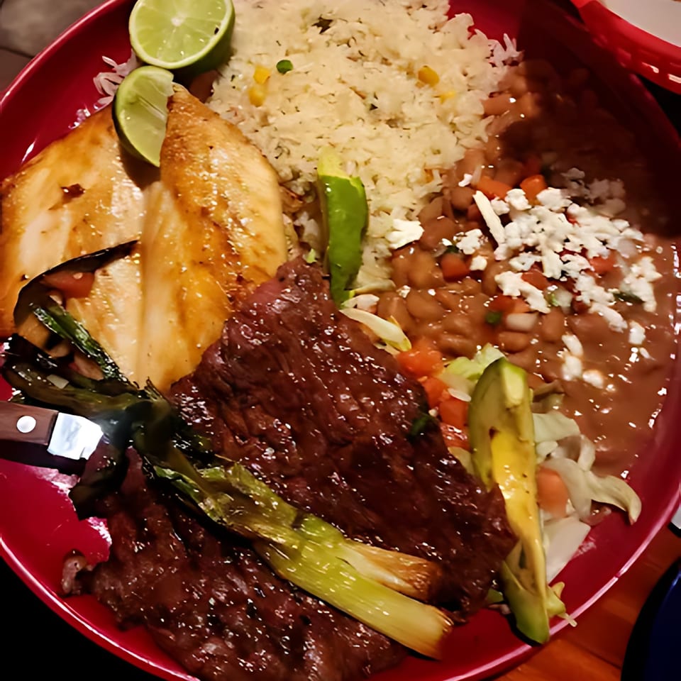 A vibrant Casa Vallarta Mexican dish with steak, chicken, rice, beans, avocado, and lime on a red plate.
